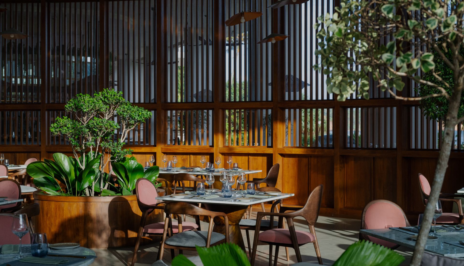 Tables laid with glasses and pink chairs among potted plants and a tree, before a wall of timber screens